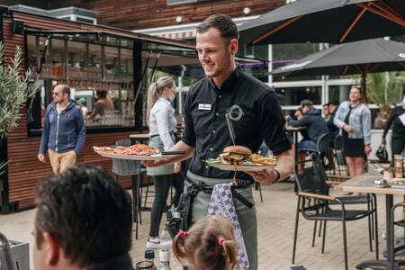 Ein Kellner bringt das Abendessen auf die Terrasse des Roompot Vakantiepark Kijkduin